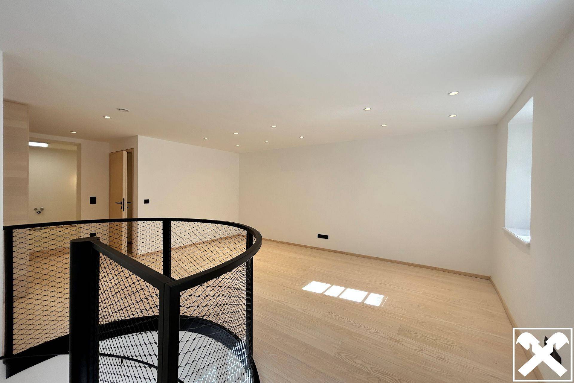 Geräumiger, heller Raum mit Holzboden, Einbaustrahlern und einer eleganten Wendeltreppe mit Metallgeländer.