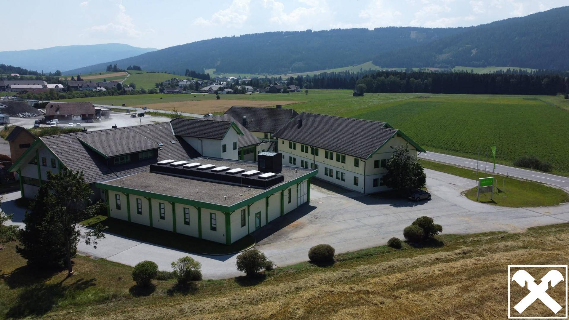 Luftaufnahme eines Gewerbekomplexes mit umliegenden Feldern und Bergen im Hintergrund unter blauem Himmel.