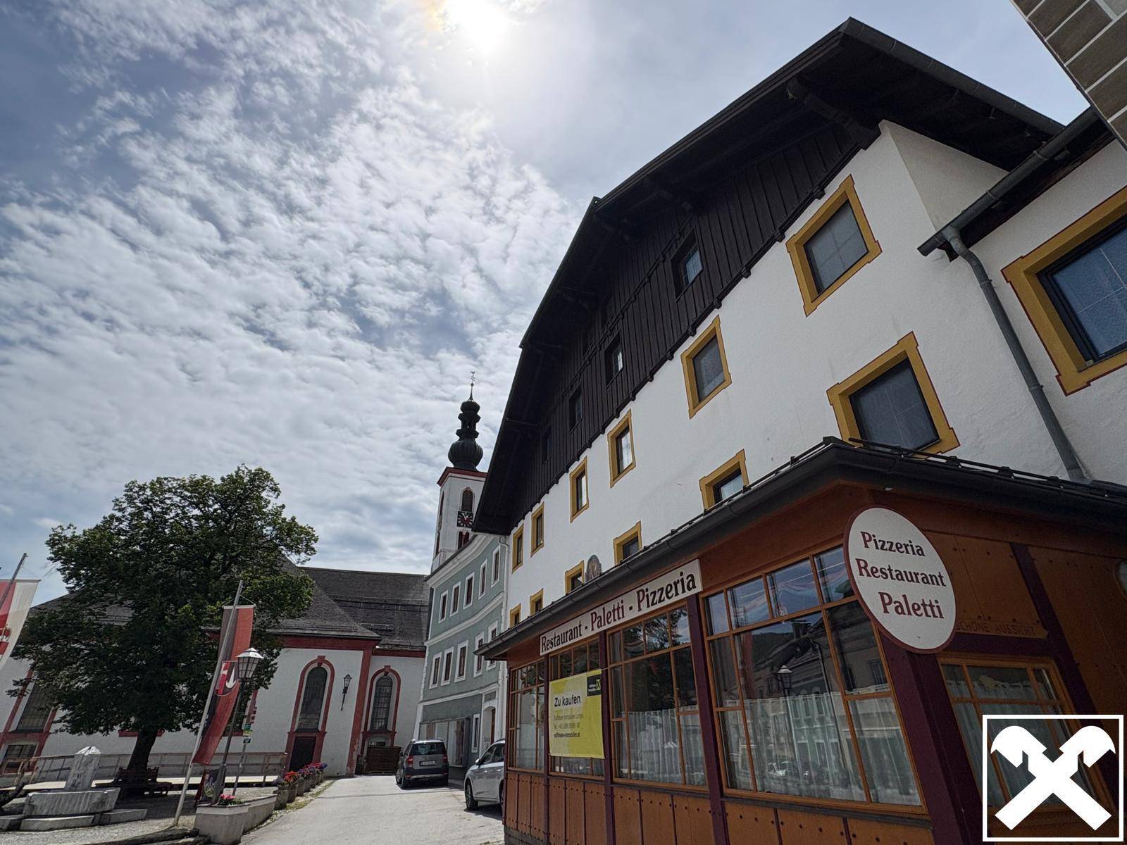 Historisches Gebäude mit Restaurant und Pizzeria, umgeben von traditioneller Architektur unter blauem Himmel.