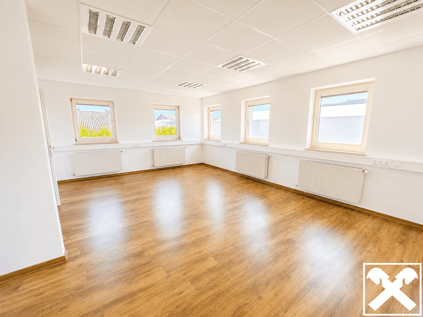 Leeres Büro mit hellem Laminatboden, weißen Wänden und mehreren Fenstern, die für natürliches Licht sorgen.