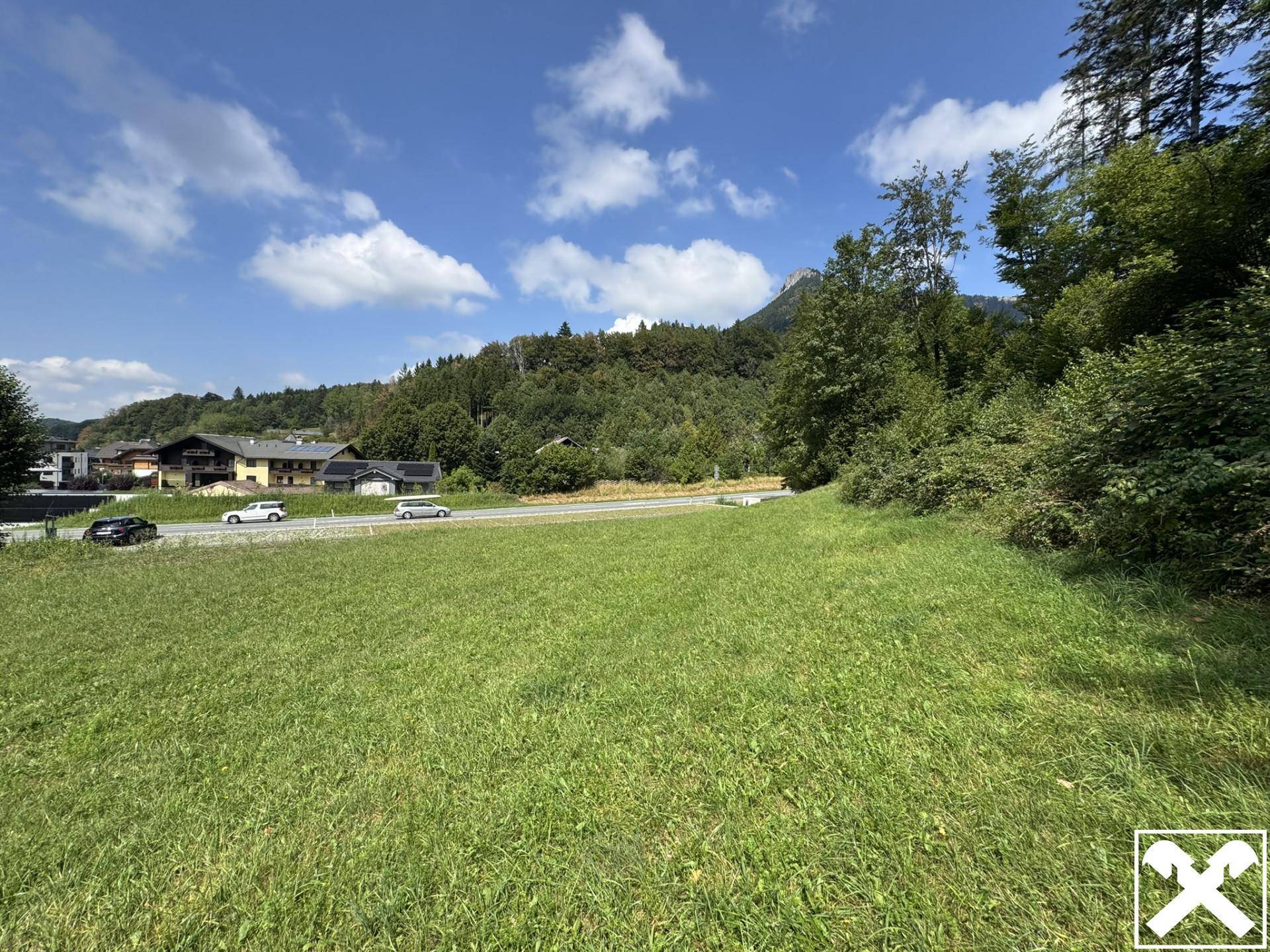 Ausgedehnte Wiese mit Blick auf eine Straße, Häuser und bewaldete Berge unter blauem Himmel.