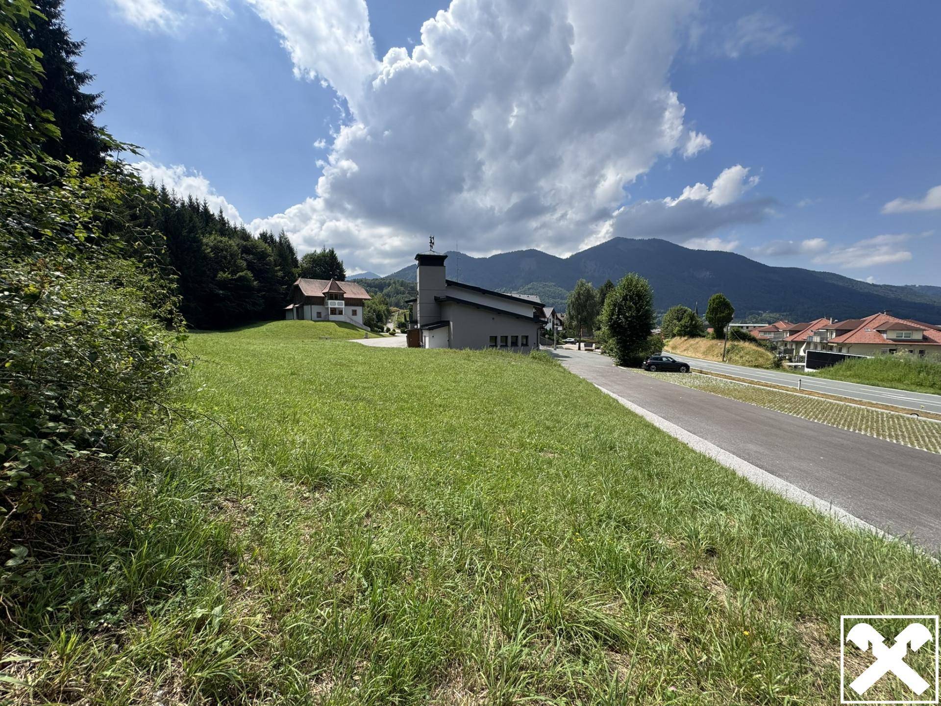 Grüne Hanglage mit Blick auf ein modernes Gebäude, eine Straße und die umliegende Bergkulisse.