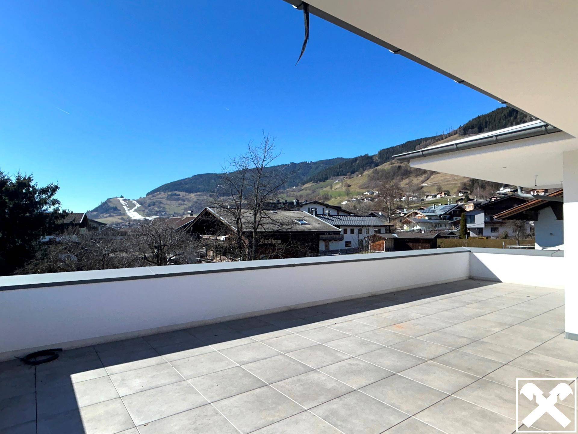 Geräumige, leere Terrasse mit Fliesenboden und Ausblick auf die umliegende Berglandschaft und das Dorf.