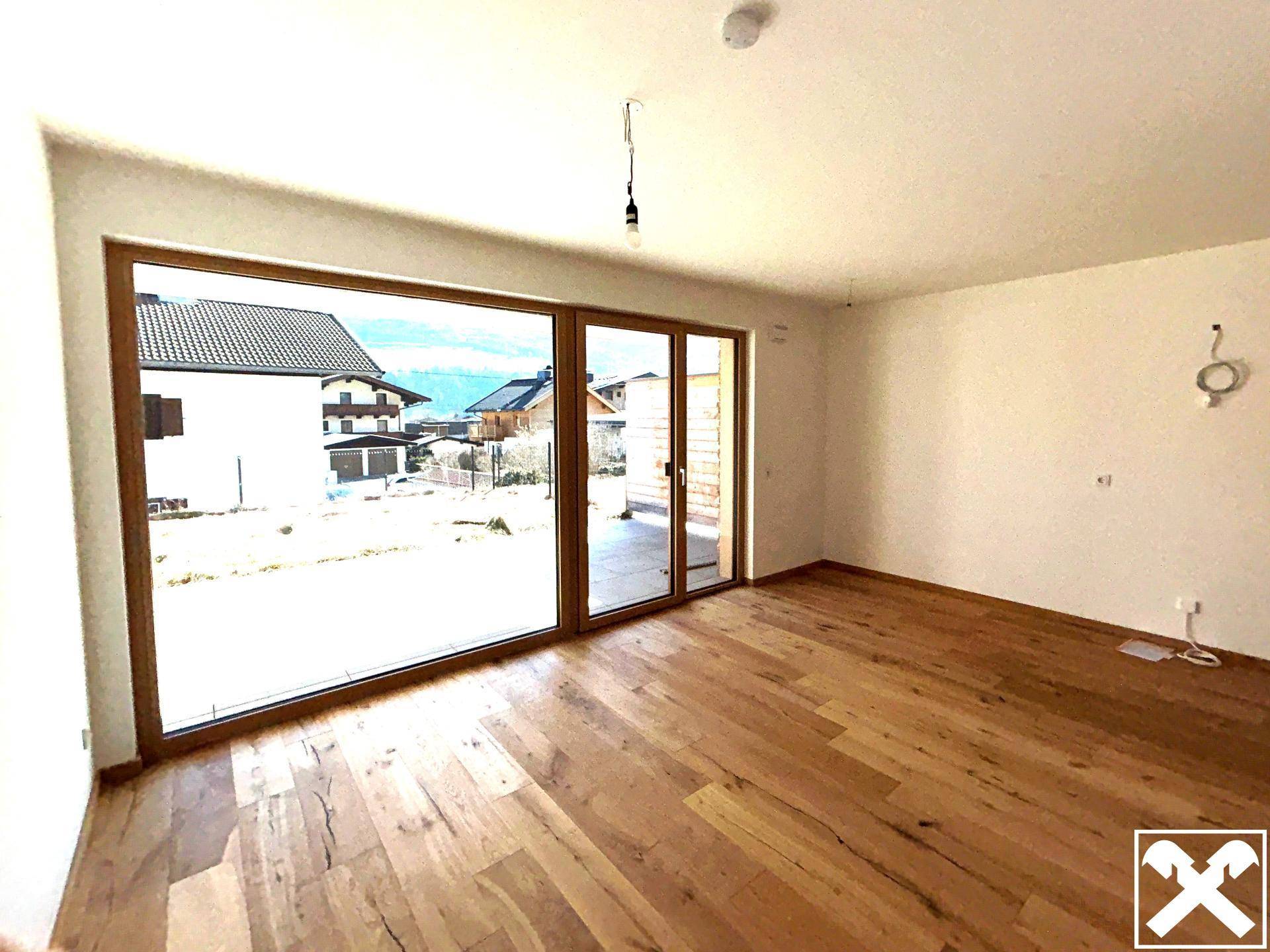 Heller Wohnraum mit großen Fenstern, Holzboden und Zugang zum Außenbereich, im Neubauzustand.