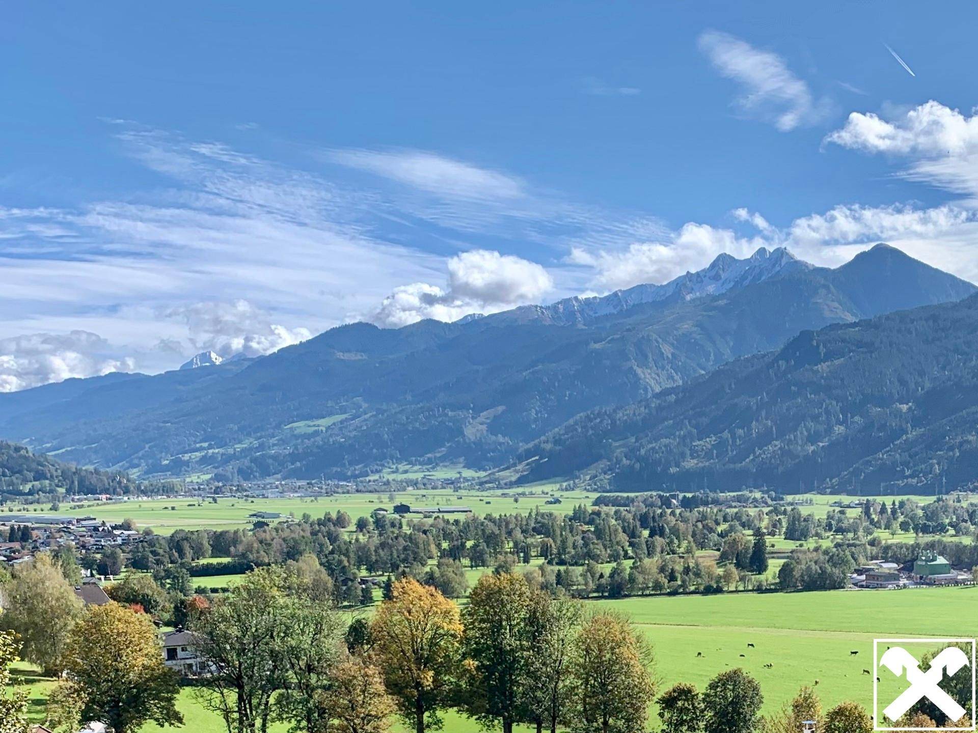Weitläufiger Blick über ein grünes Tal mit bewaldeten Hügeln und einer beeindruckenden Bergkette am Horizont.