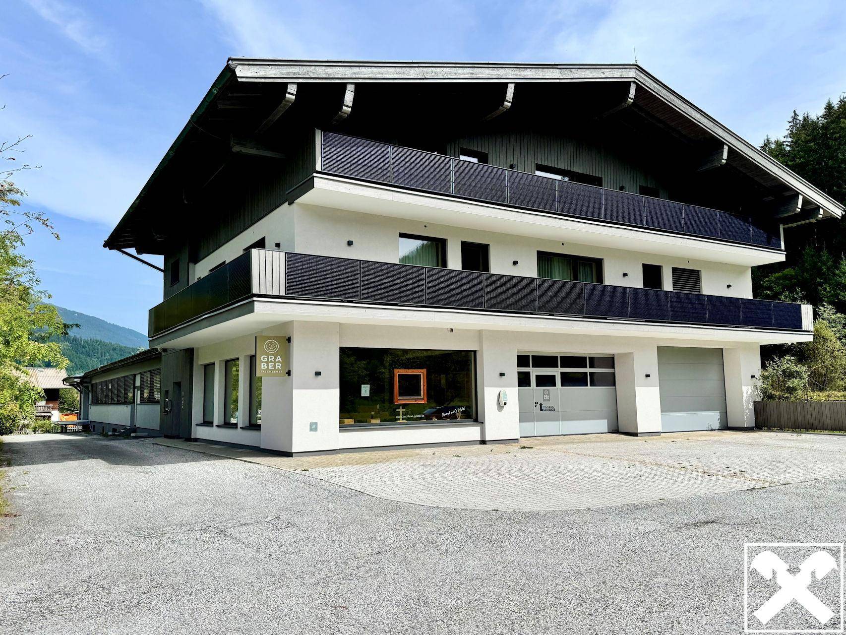 Modernes Geschäftsgebäude mit großen Fenstern, Balkonen und Solarpaneelen auf dem Dach in alpiner Landschaft.