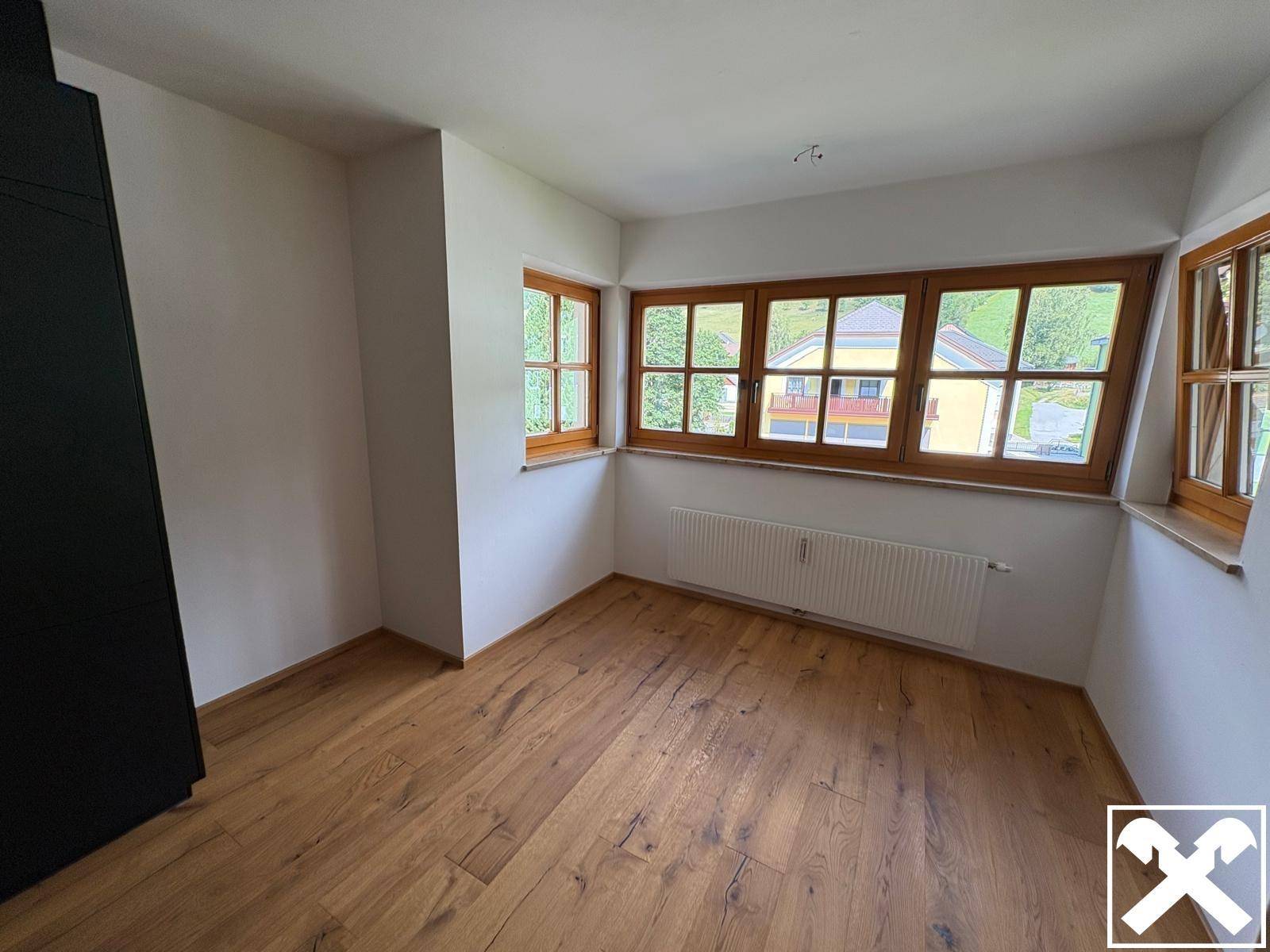 Helles Zimmer mit Holzboden und mehreren Fenstern, die einen grünen Ausblick bieten.