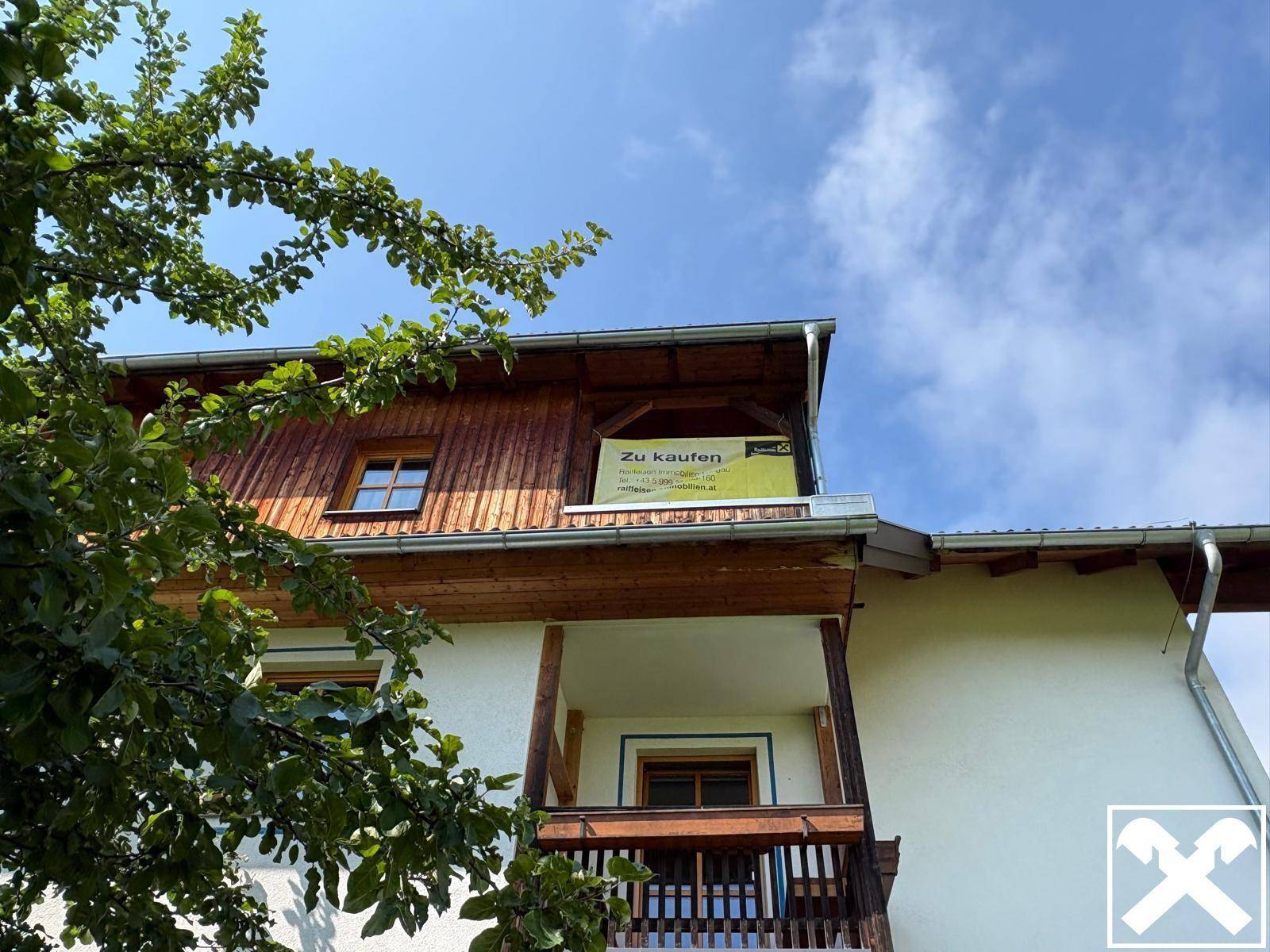 Ansicht des Gebäudes mit Holzverkleidung und einem "Zu kaufen"-Schild unter blauem Himmel.