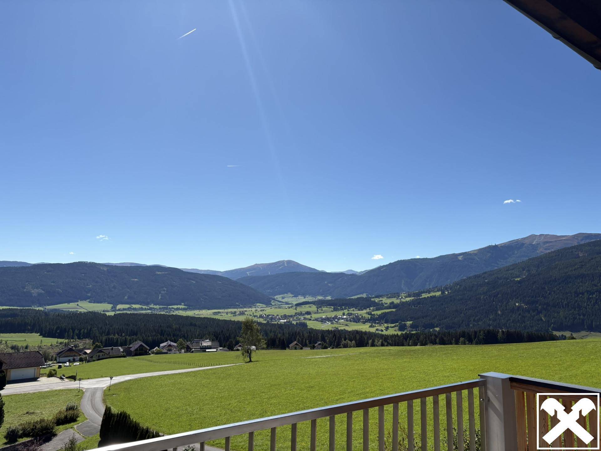 Weitläufiger Panoramablick vom Balkon über grüne Wiesen und die umliegende Berglandschaft an einem sonnigen Tag.