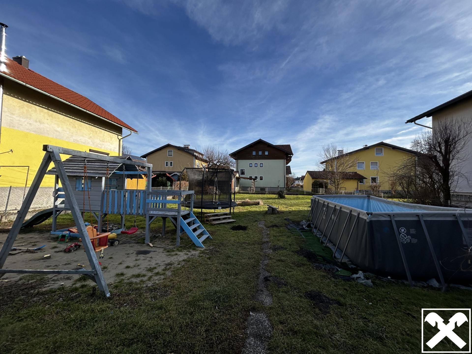 Gartenbereich mit Kinderspielplatz, Schaukel und einem Aufstellpool unter blauem Himmel.