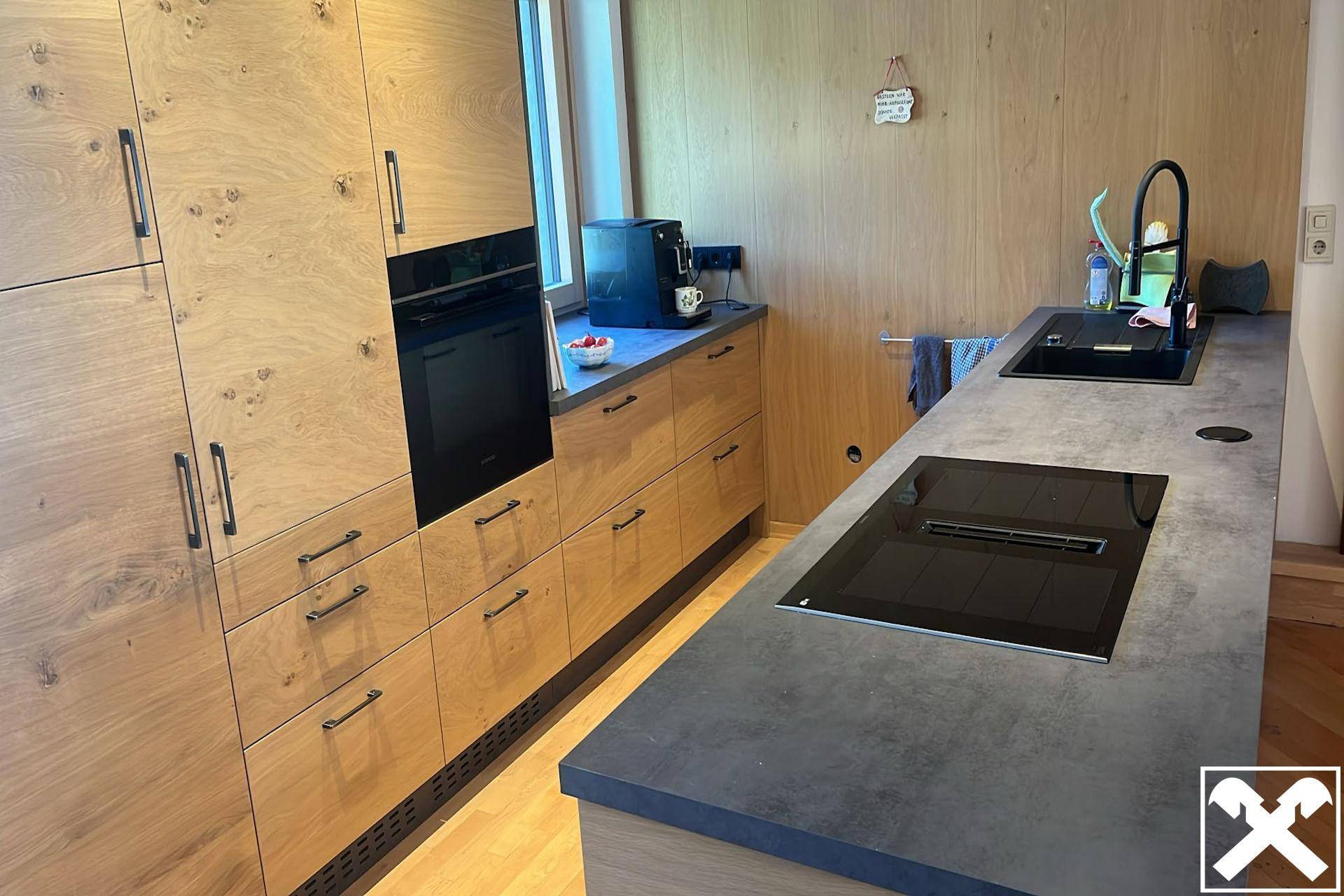 Moderne Küche mit hellen Holzfronten, dunklen Arbeitsplatten und integrierten Geräten. Ein Fenster sorgt für natürliches Licht.