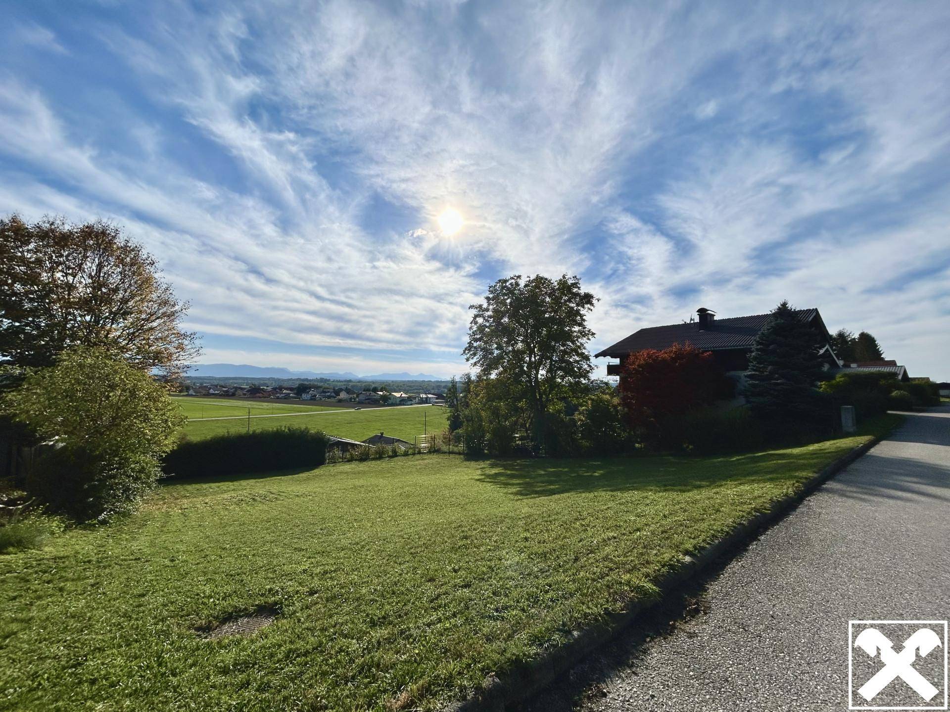 Sonnenbeschienener Blick auf ein Wohnhaus an einer Straße mit weitläufigen Feldern und Bergen im Hintergrund.