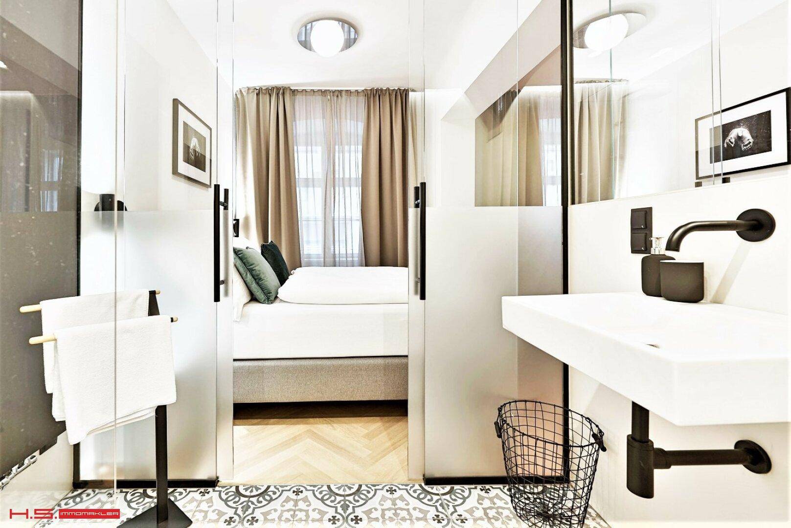 Modernes Badezimmer mit Waschbecken, Spiegel und Blick ins Schlafzimmer durch eine Glasabtrennung.