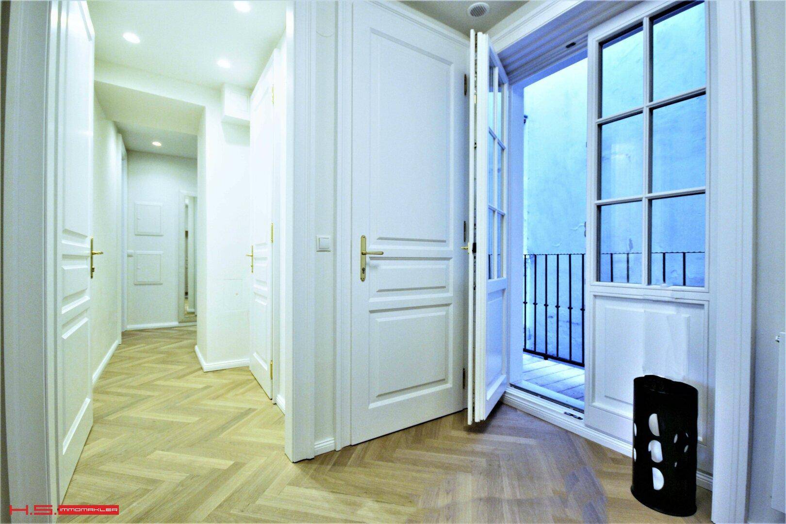 Heller Flur mit Fischgrätparkett und Zugang zu einem französischen Balkon durch eine offene Tür.