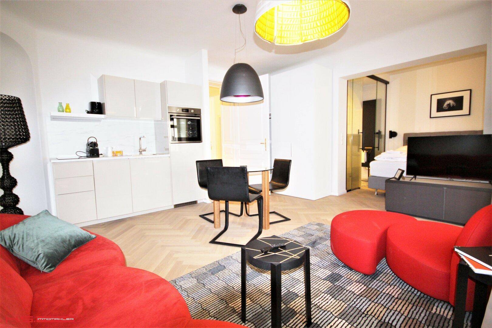 Offener Wohn-Essbereich mit rotem Sofa, Küchenzeile und Blick ins Schlafzimmer durch Glaswand.