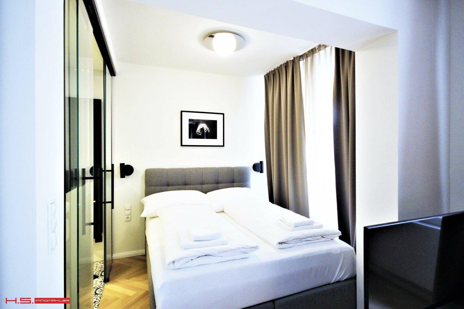 Schlafzimmer mit Doppelbett, Vorhängen und Blick ins Badezimmer durch eine Glasabtrennung.