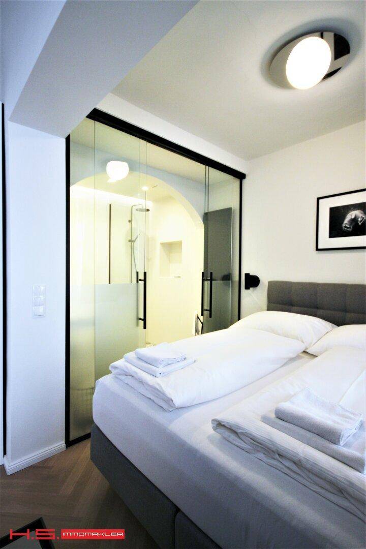 Schlafzimmer mit Doppelbett und angrenzendem Duschbereich, getrennt durch eine Glaswand.