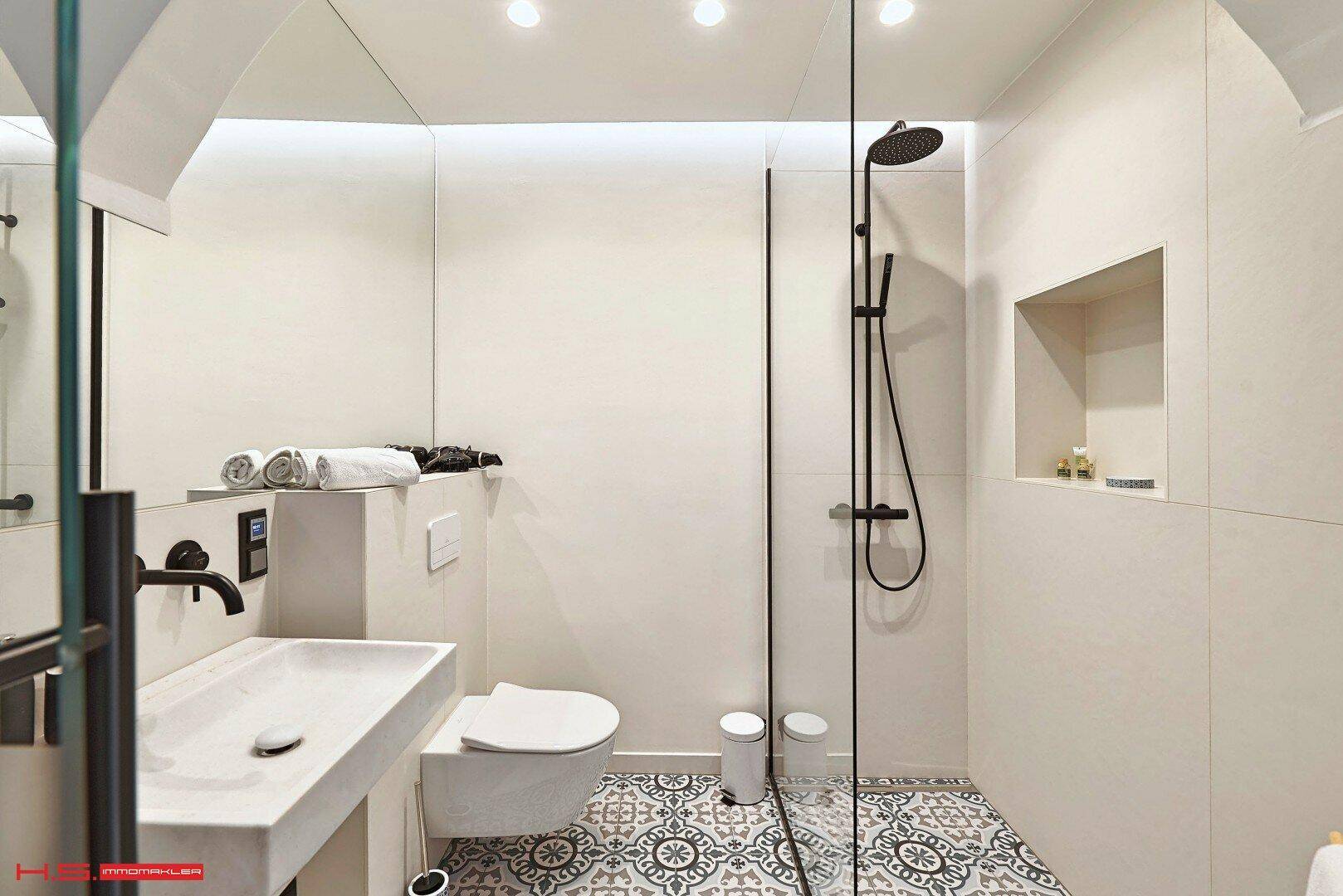 Modernes Badezimmer mit wandhängendem WC, Waschbecken und begehbarer Dusche auf gemusterten Fliesen.