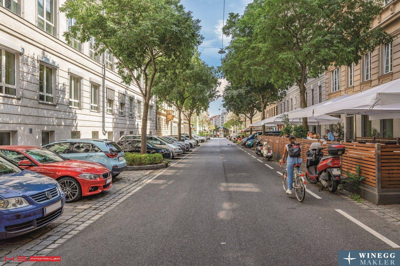 Belebte Straße mit parkenden Autos, Bäumen und einem Straßencafé vor den Wohngebäuden.