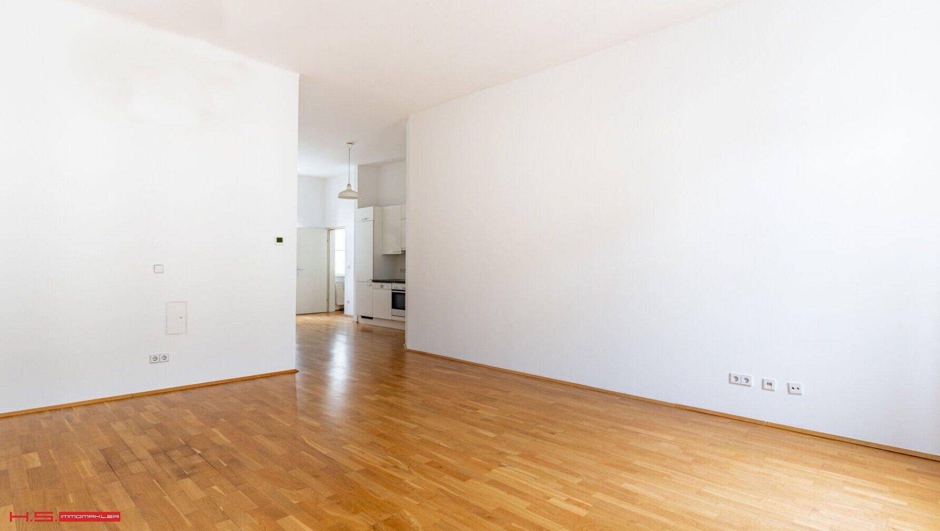 Geräumiger, leerer Wohnbereich mit hellem Parkettboden und Zugang zur Küche.
