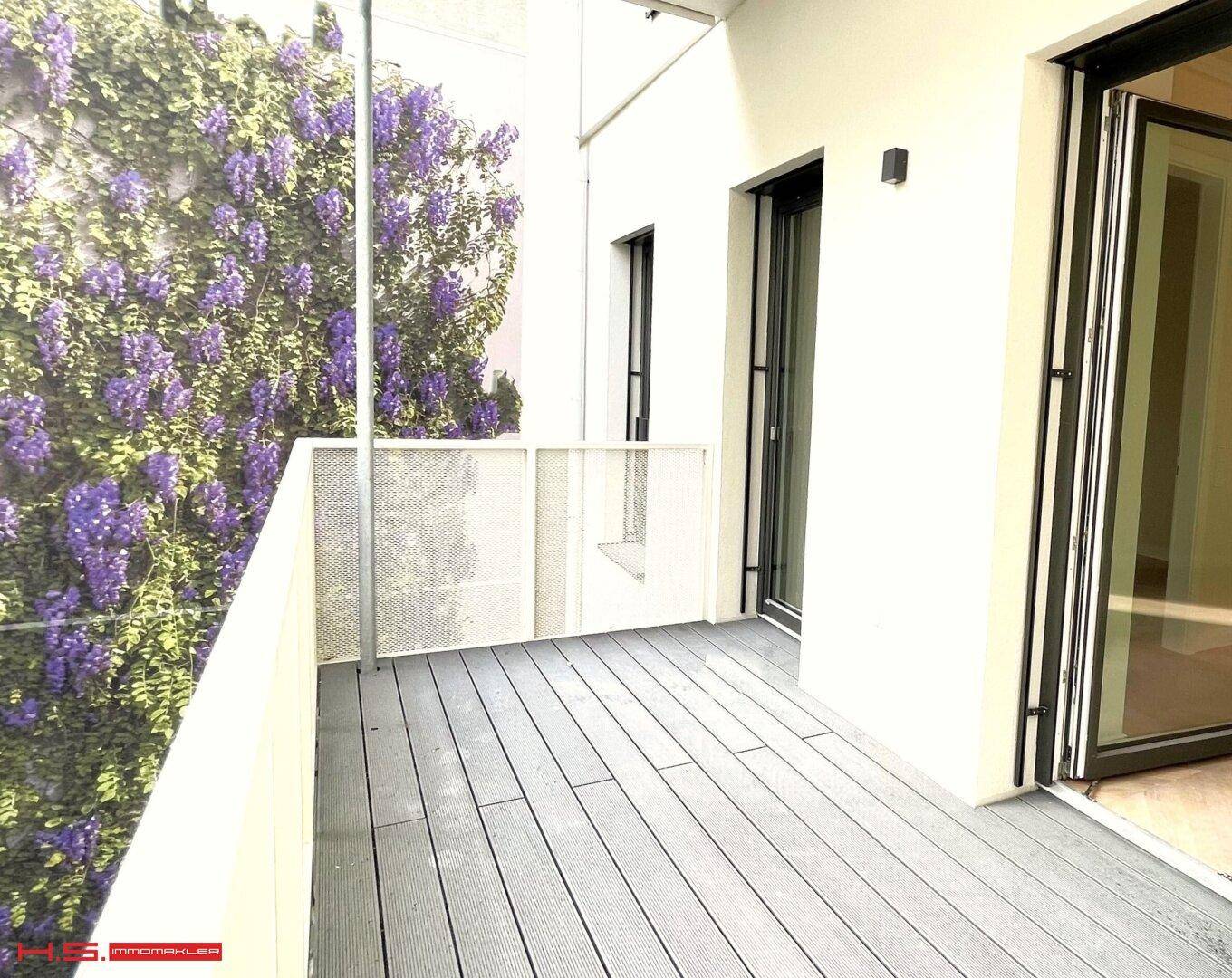 Heller Balkon mit modernem Holzboden und Zugang zum Innenbereich, umgeben von üppigem Grün.