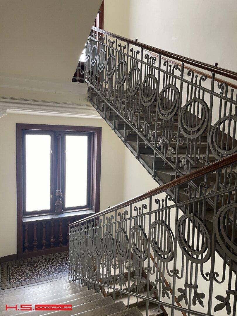 Historisches Treppenhaus mit kunstvollem Geländer aus Schmiedeeisen und großen Fenstern.