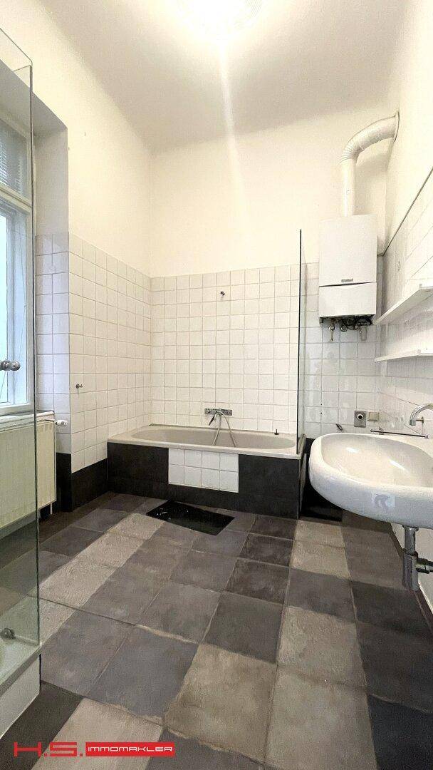 Badezimmer mit Badewanne, Waschbecken und Fenster, ausgestattet mit weißen und grauen Fliesen.