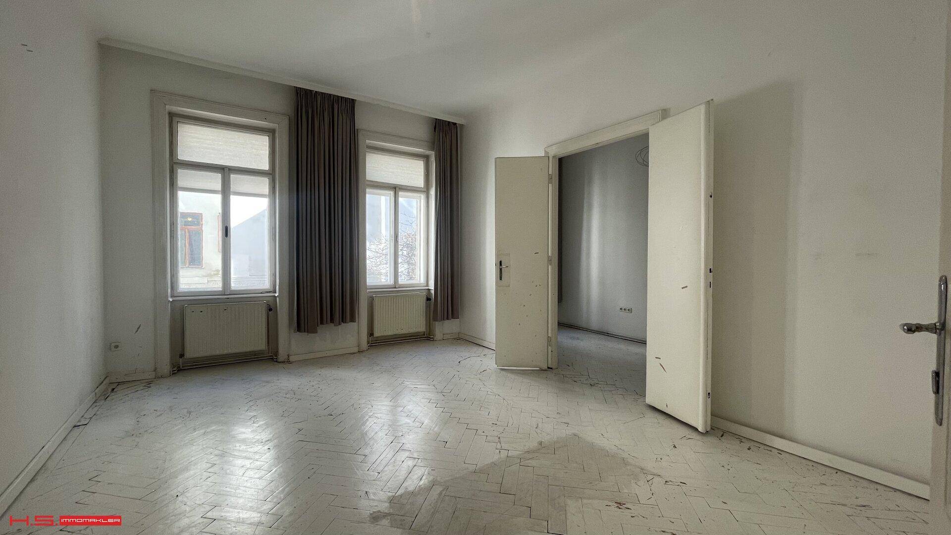 Geräumiges Zimmer mit Fischgrätparkett, hohen Fenstern und Vorhängen, ideal als Schlaf- oder Wohnzimmer.