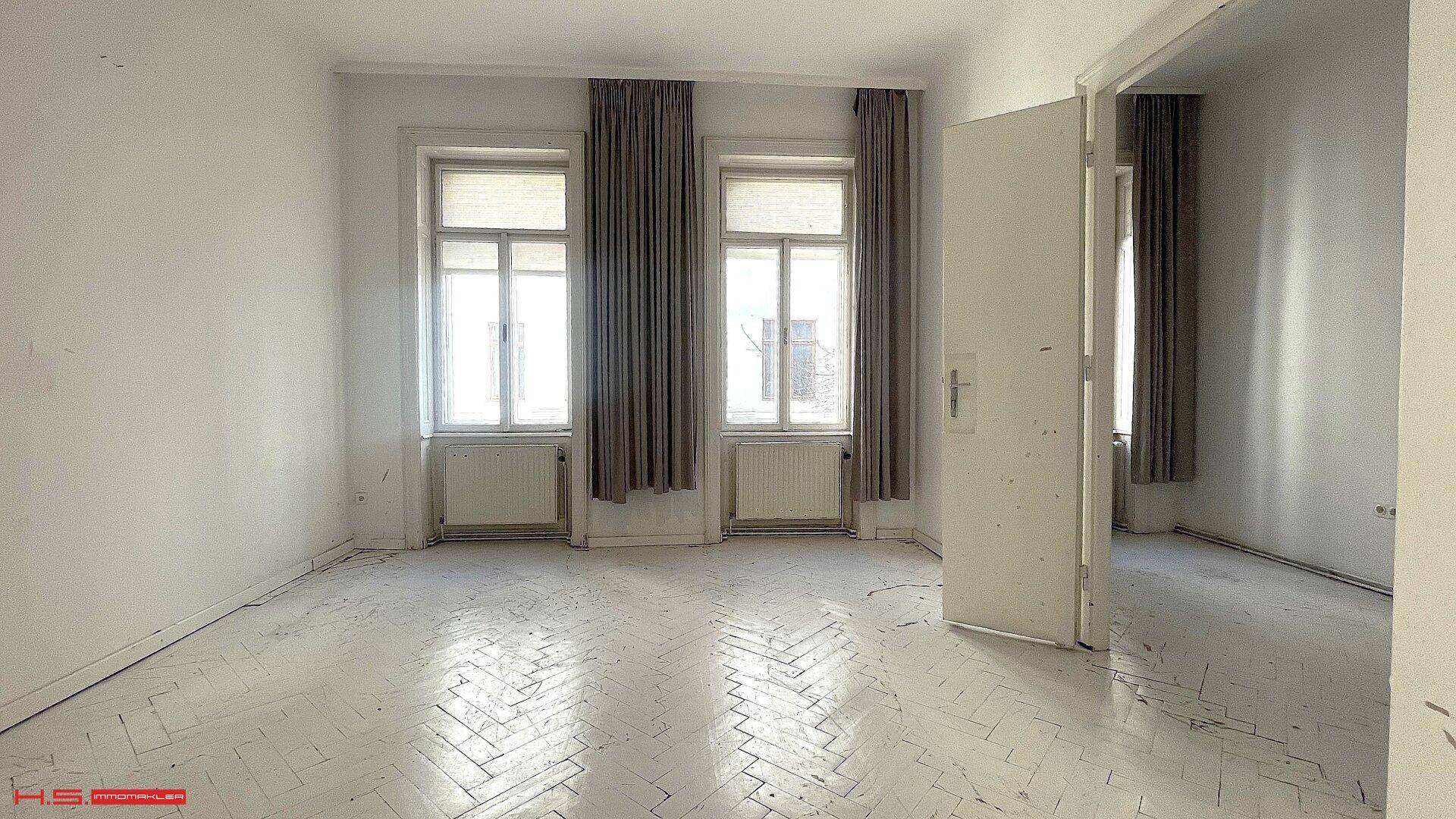 Helles Zimmer mit zwei hohen Fenstern, Vorhängen und Fischgrätparkett, ideal als Schlafzimmer.