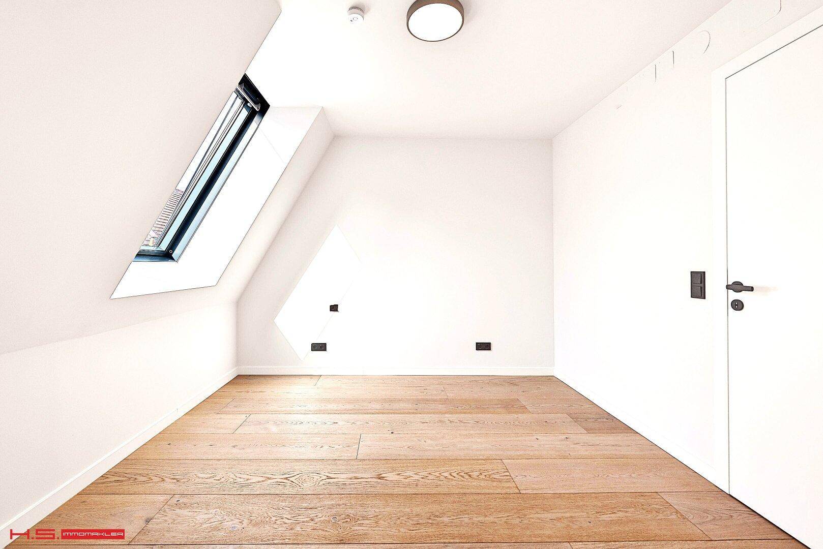 Helles Zimmer im Dachgeschoss mit Holzboden, schrägen Wänden und einem großen Dachfenster.
