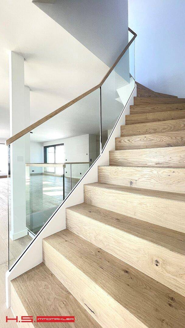 Moderne Holztreppe mit Glasgeländer und hellem Holzparkett in einem lichtdurchfluteten Bereich.