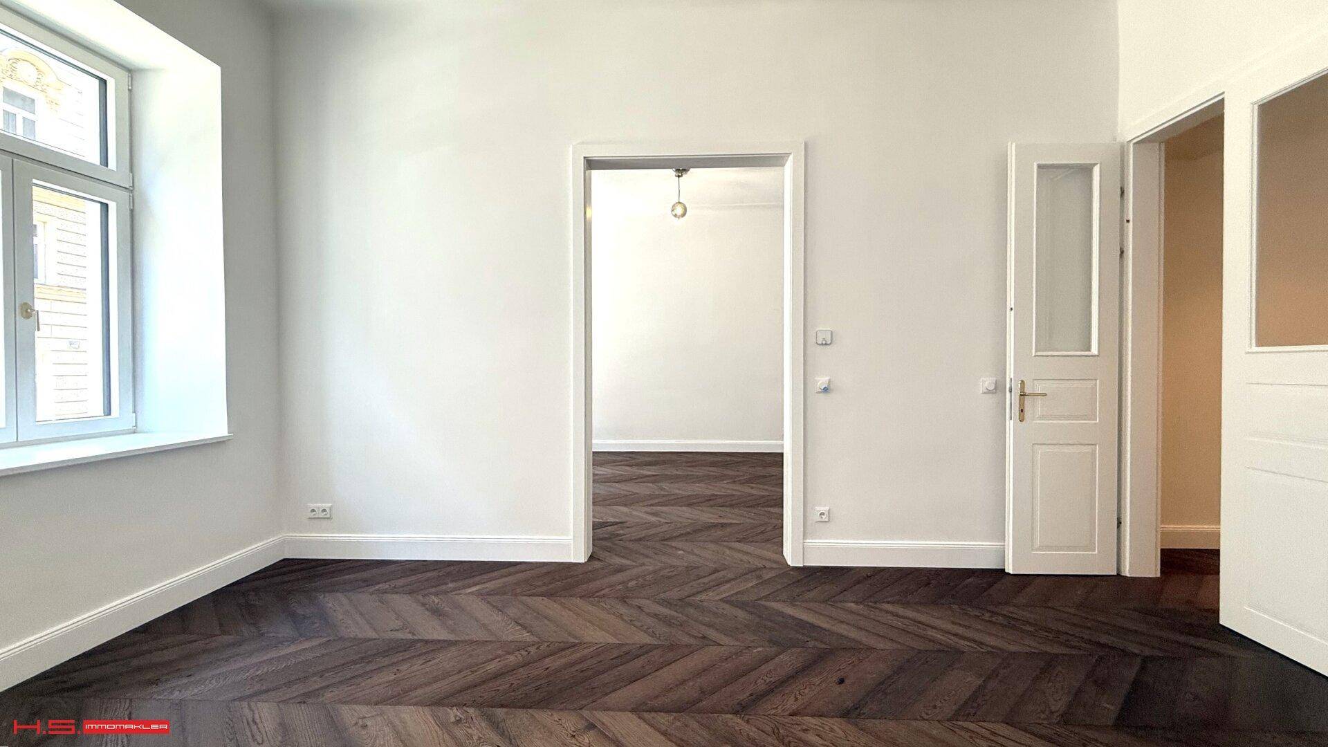 Geräumiges Zimmer mit elegantem Fischgrätparkett und Zugang zu weiteren Räumen durch eine offene Tür.