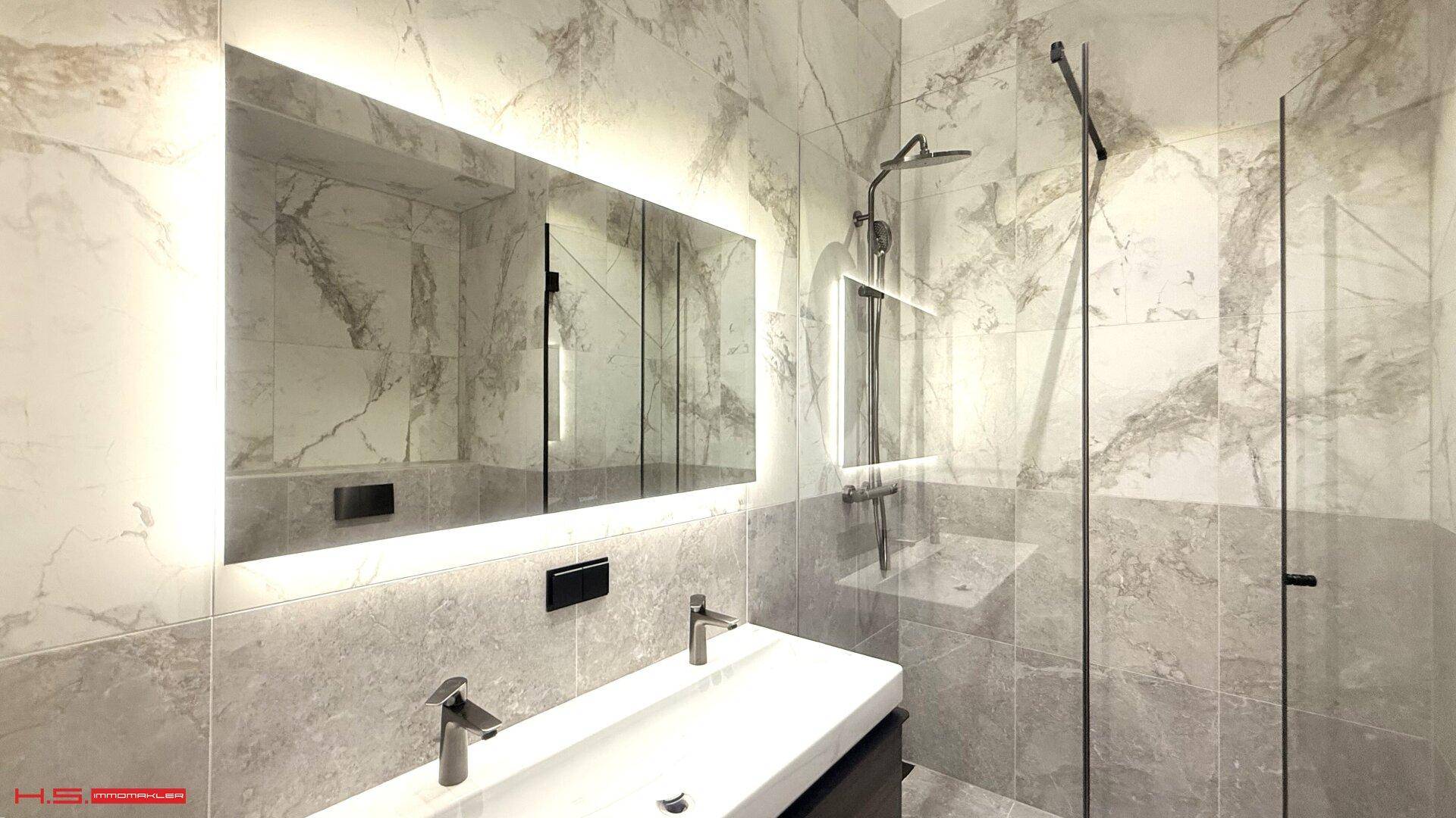Modernes Badezimmer mit Doppelwaschbecken, beleuchtetem Spiegel und begehbarer Dusche, gefliest mit Marmoroptik.