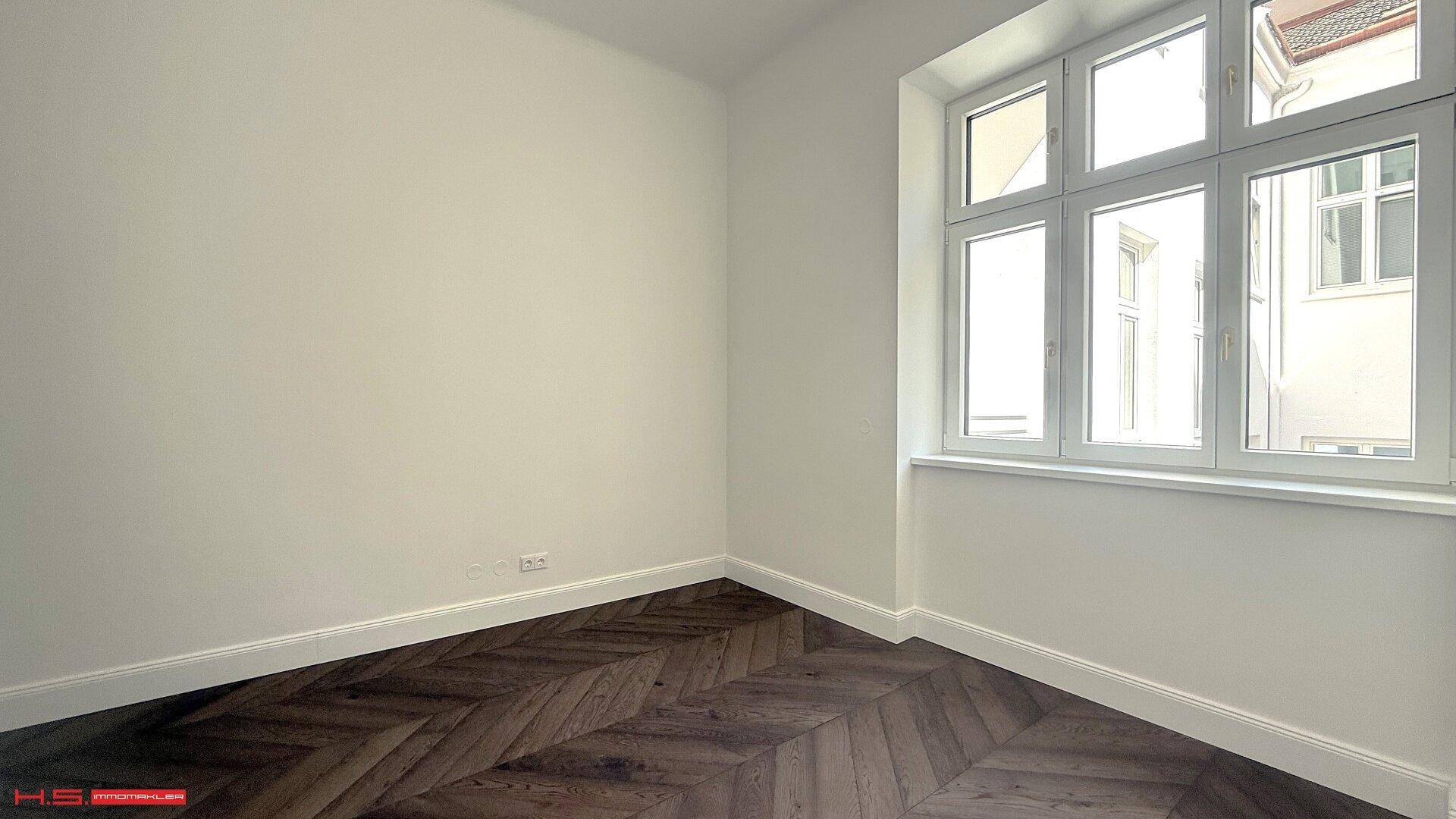 Helles Schlafzimmer mit dunklem Fischgrätparkett und einem großen Fenster, das den Raum erhellt.