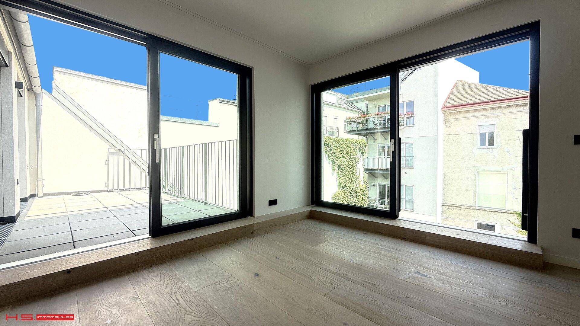 Heller Raum mit Holzboden, großen Schiebetüren zur Terrasse und Blick auf begrünte Gebäude.