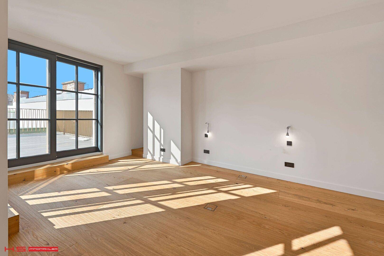 Heller Raum mit Holzboden und großen Fenstertüren, die viel Sonnenlicht hereinlassen und Zugang nach draußen bieten.