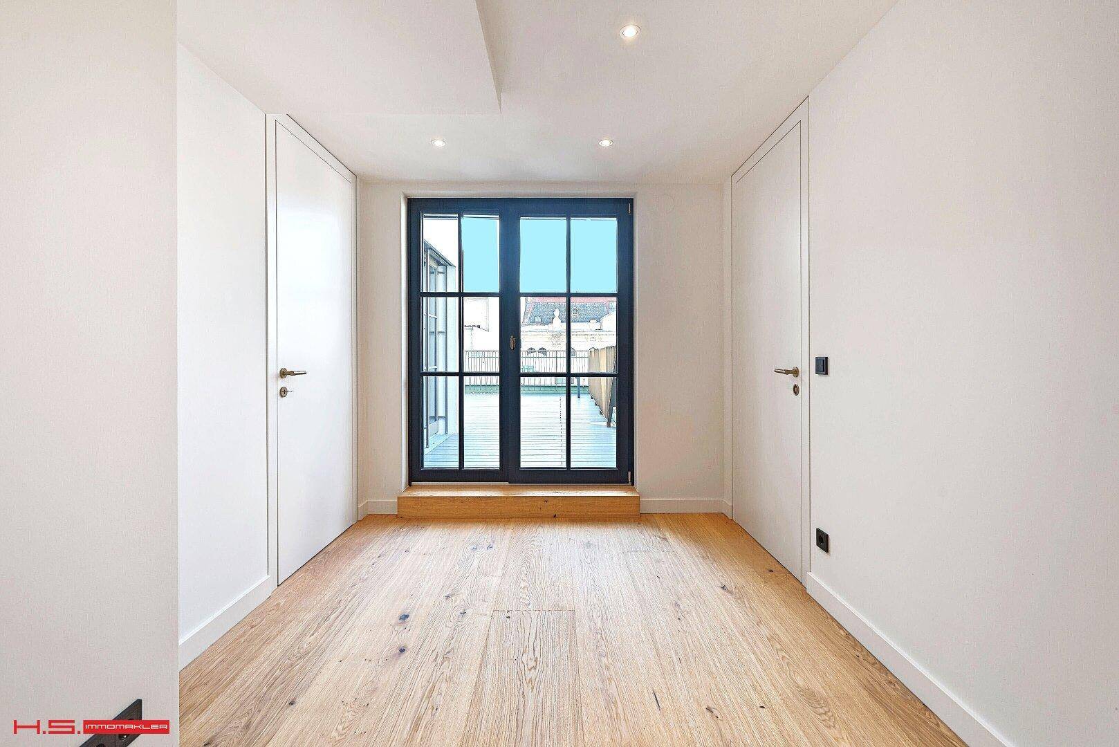 Helles Zimmer mit Holzboden und großen Fenstertüren, die Zugang zu einem Außenbereich ermöglichen.
