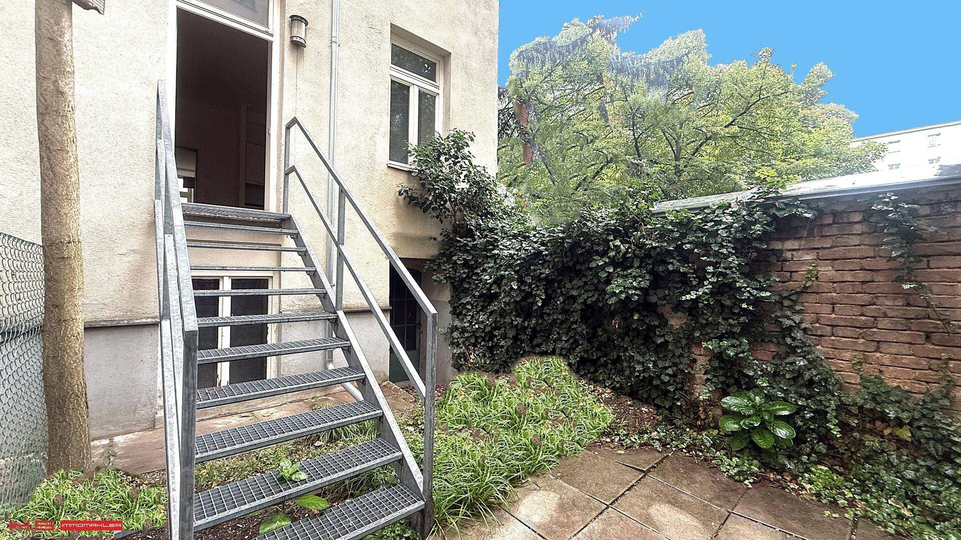 Außenbereich mit Metalltreppe zum Eingang und bewachsenen Ziegelmauern.