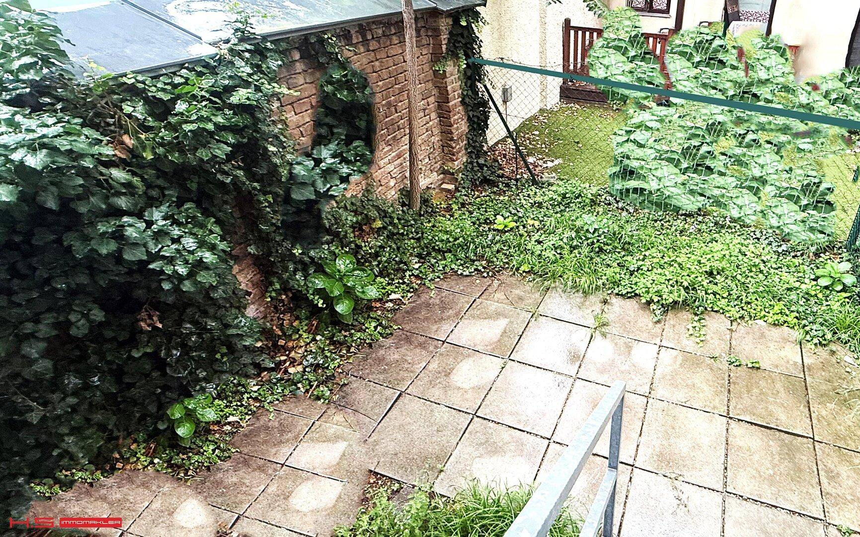 Kleiner, verwilderter Gartenbereich mit Steinplatten und viel Grünbewuchs.