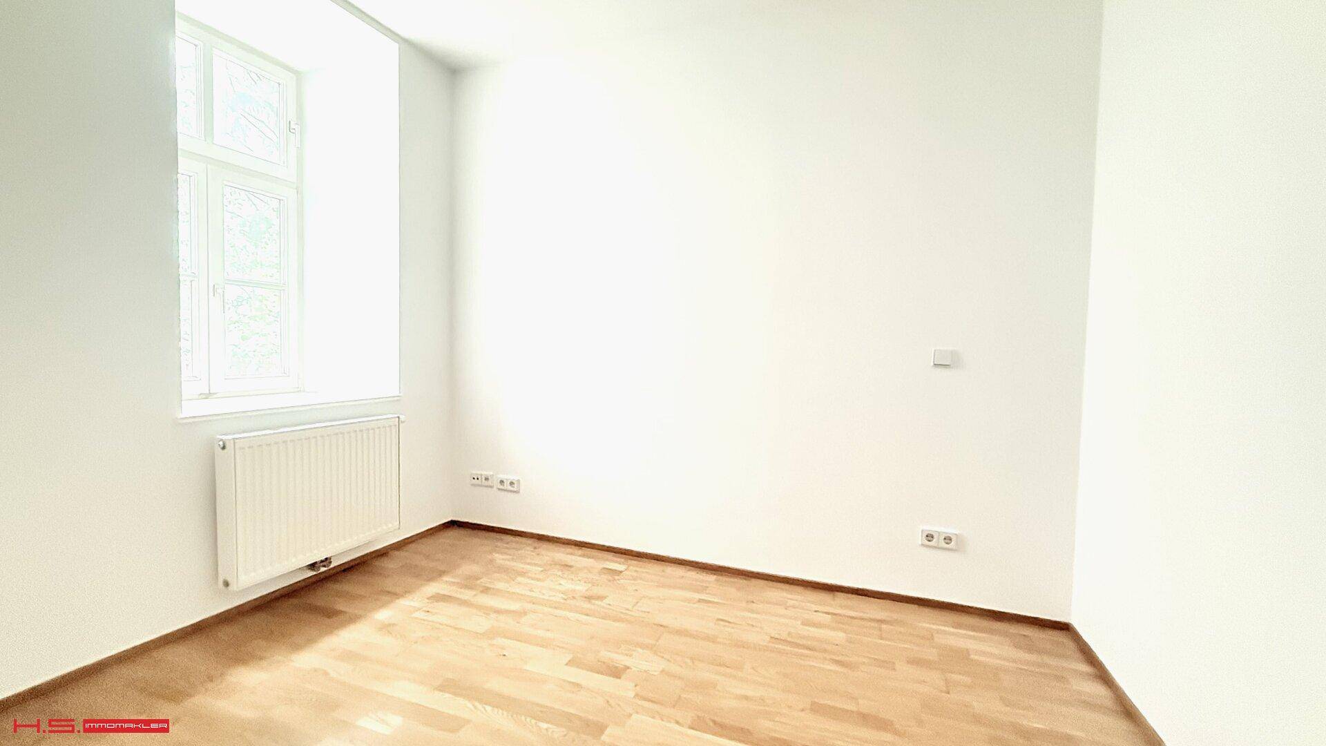 Helles Zimmer mit einem Fenster, weiß gestrichenen Wänden und Parkettboden.