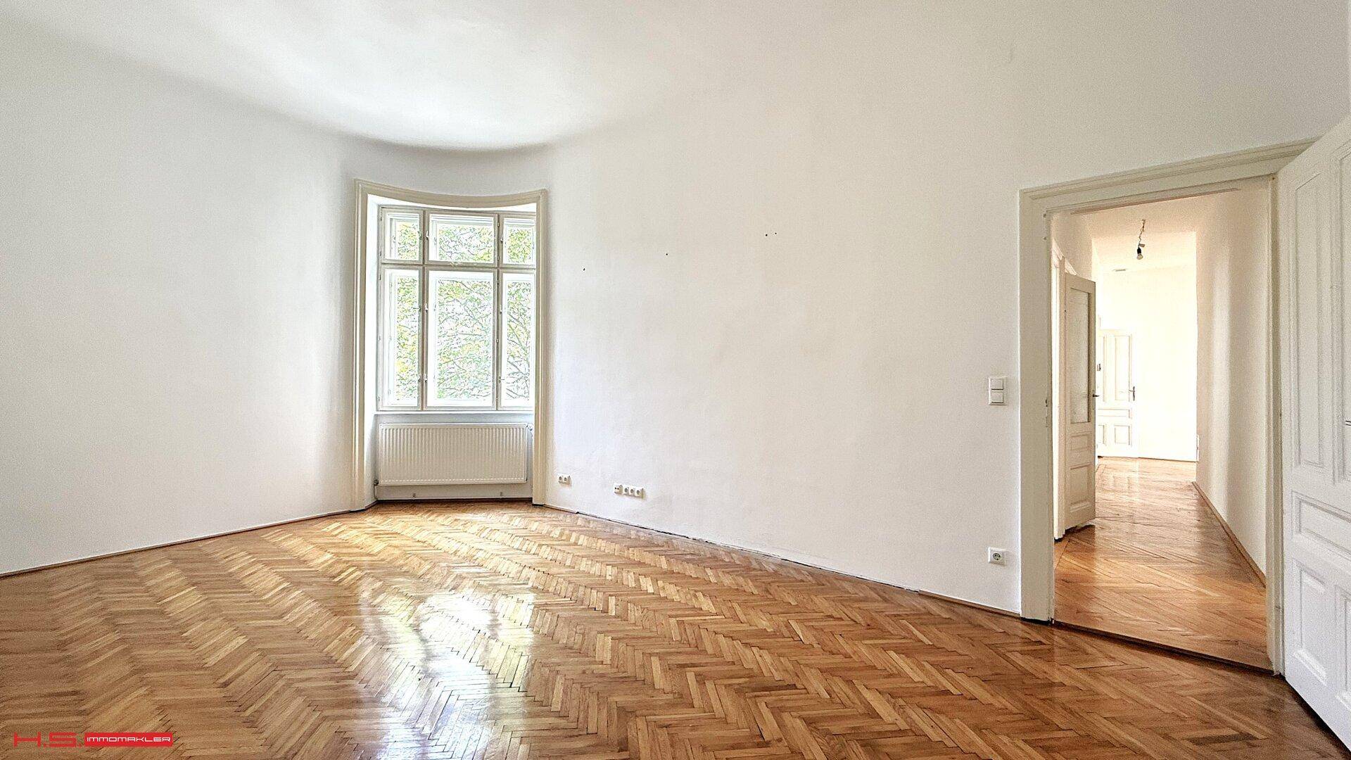 Helles, leeres Zimmer mit Fischgrätparkett und einem großen Fenster mit Blick ins Grüne.