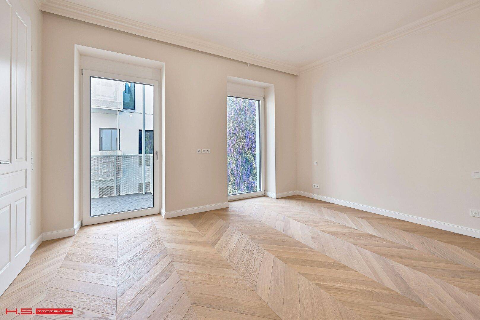 Geräumiger Raum mit hellem Fischgrätparkett und zwei hohen Fenstertüren, die viel Tageslicht hereinlassen.