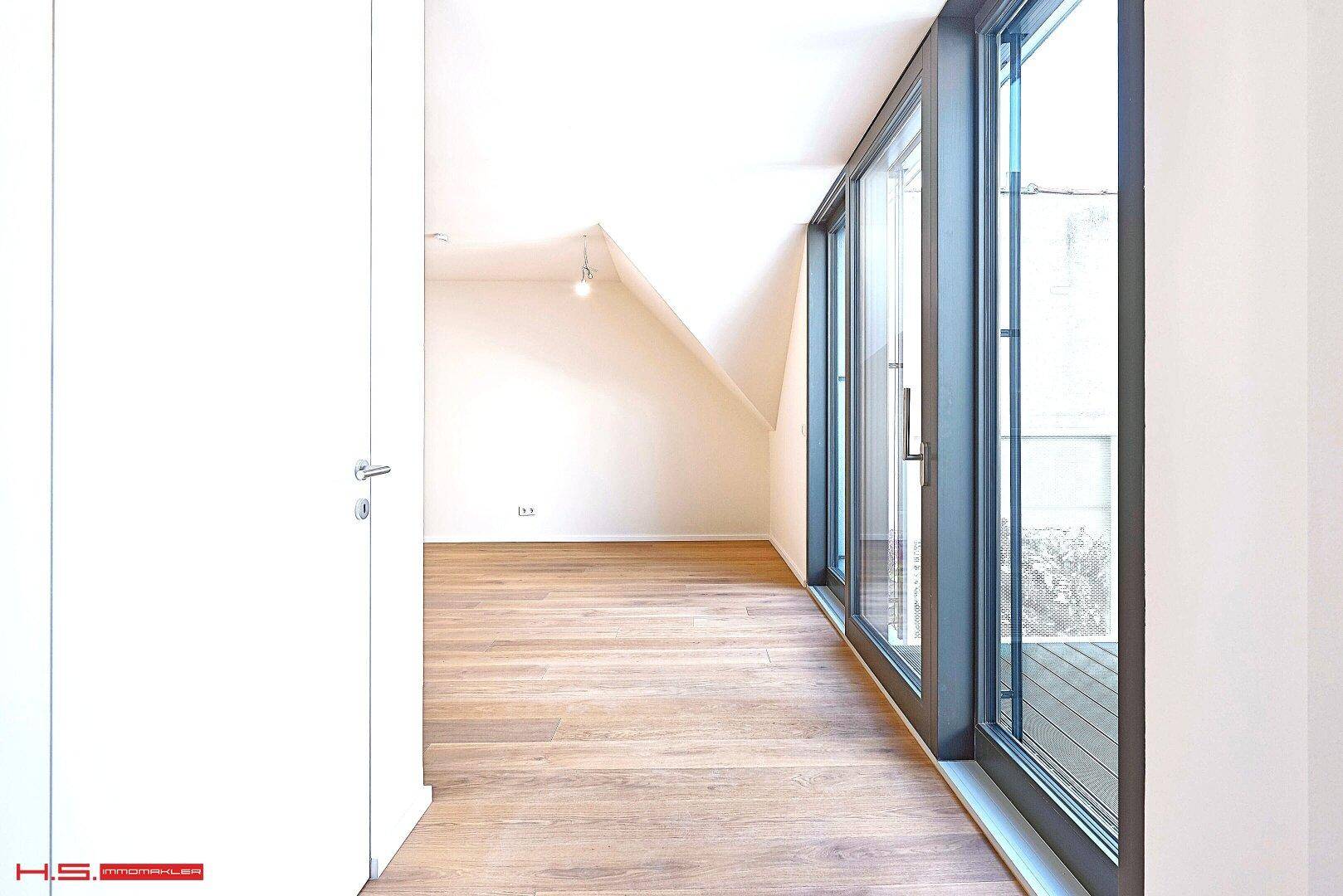 Heller Raum mit Holzboden und großen Fenstern, die den Blick nach draußen freigeben.