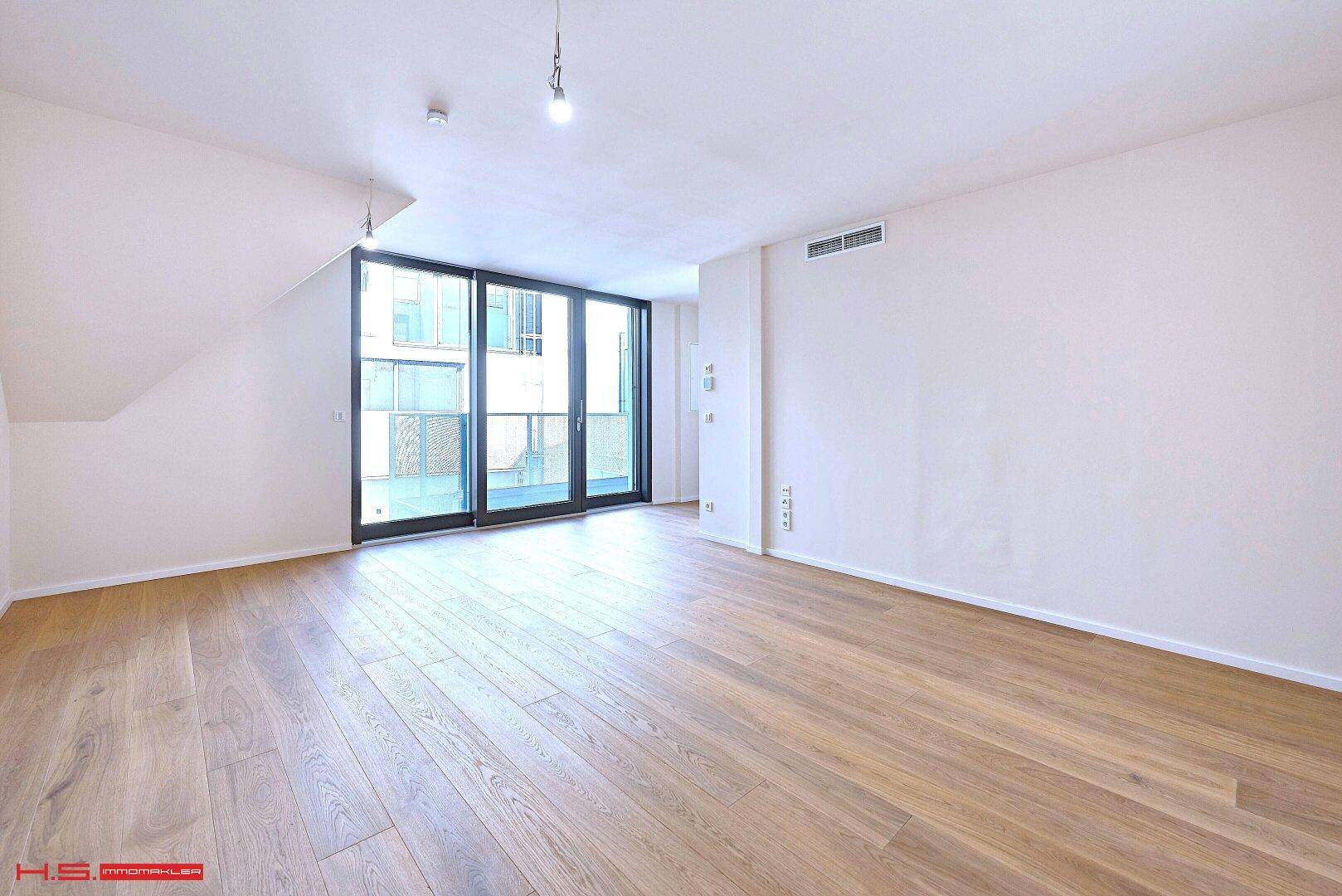 Heller, leerer Raum mit Holzboden und großen Fenstern, die den Zugang zum Balkon ermöglichen.