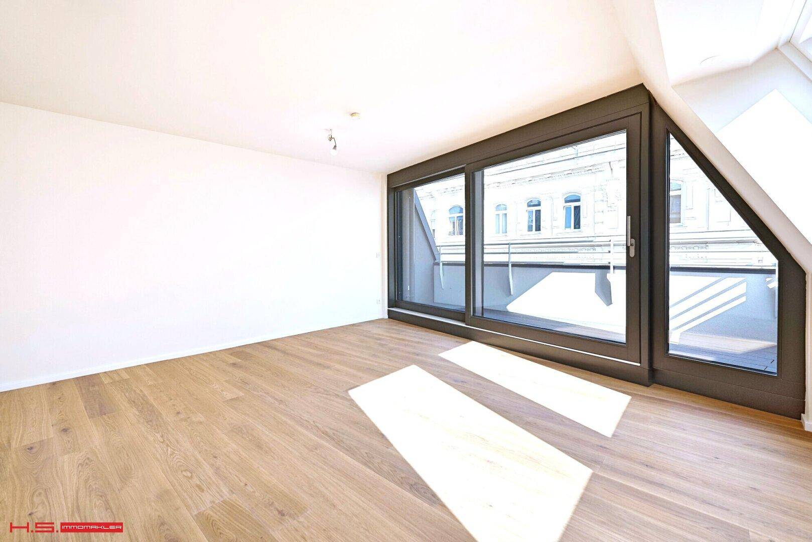 Geräumiger, heller Raum mit Holzboden, Schiebetüren zum Balkon und schräger Decke mit Dachfenster.
