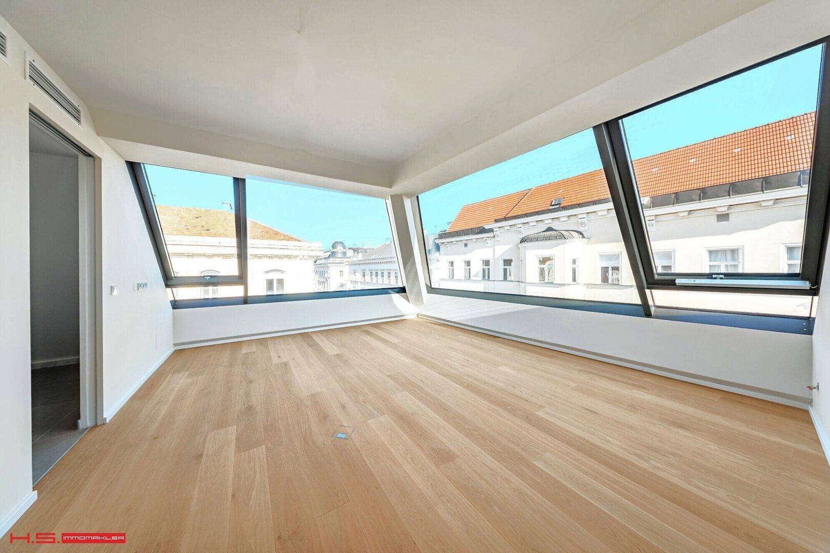 Lichtdurchfluteter Raum mit Holzboden und großen, schrägen Fenstern, die einen urbanen Blick bieten.