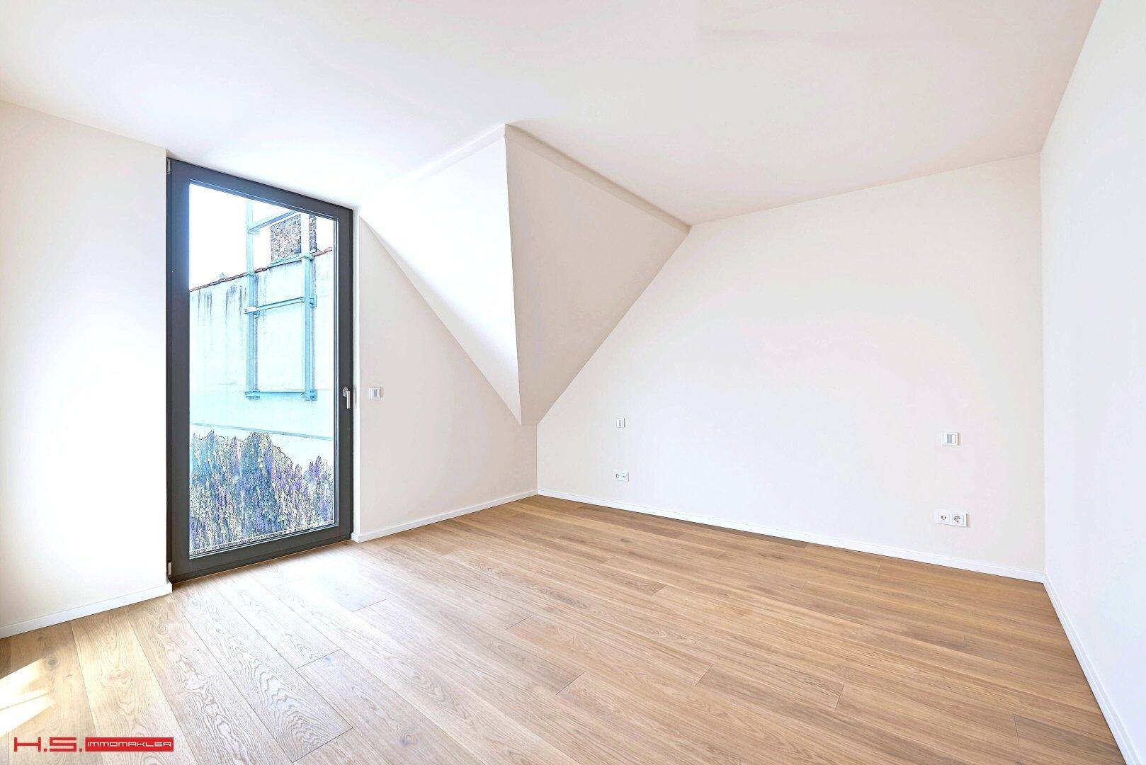 Leeres Zimmer mit Holzboden, Dachschräge und einem hohen Fenster, das viel Licht spendet.
