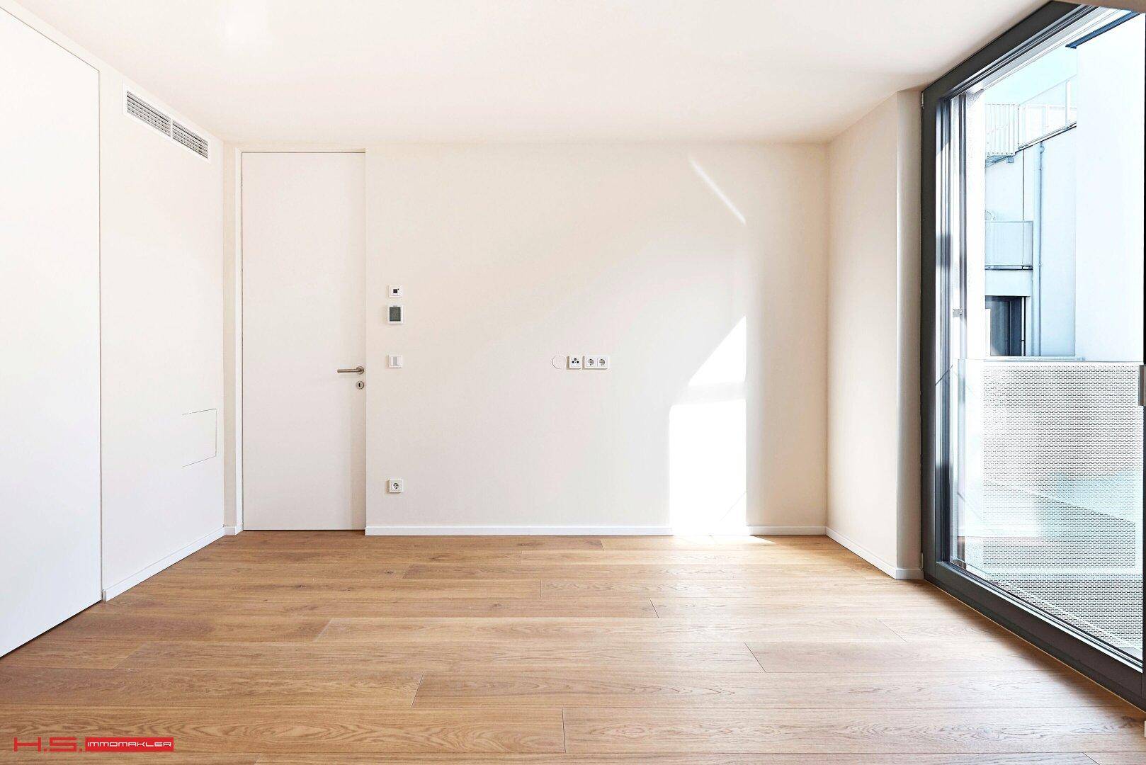 Leeres Zimmer mit Holzboden und Zugang zu einem Balkon durch eine große Glastür.