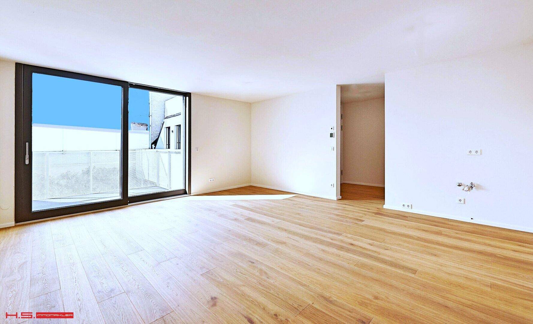 Geräumiger, leerer Wohnbereich mit großen Schiebetüren zum Balkon und hellem Parkettboden.
