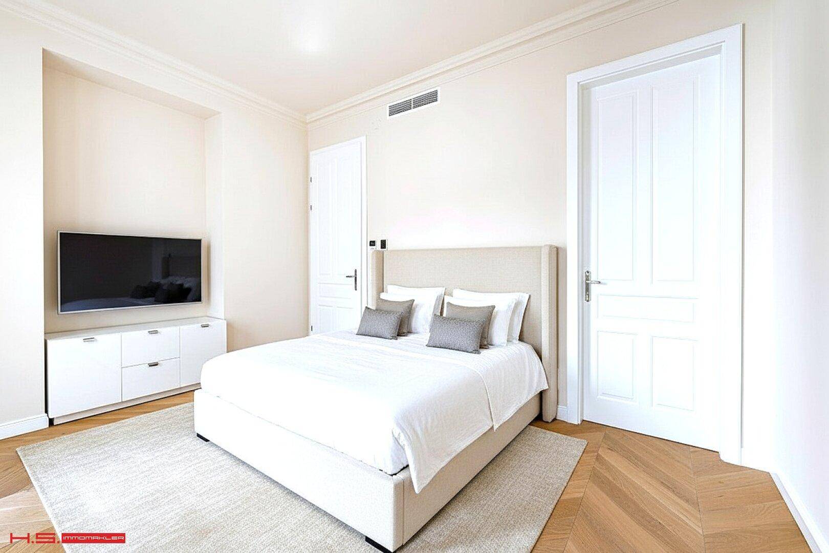 Helles Schlafzimmer mit Doppelbett, Fernseher und elegantem Fischgrätparkett.